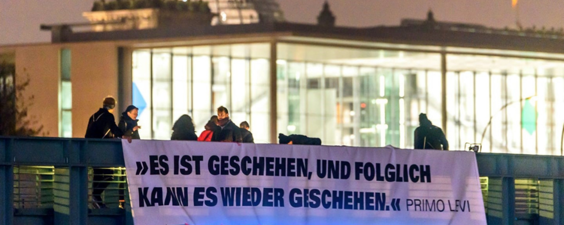 Man erkennt eine Brücke im Vordergrund, an der Personen ein Banner mit der Aufschrift "Es ist geschehen, und folglich kann es wieder geschehen." (Primo Levi) angebracht haben. Im Hintergrund ist unscharf das Reichstagsgebäude mit der Glaskuppel zu erkennen.