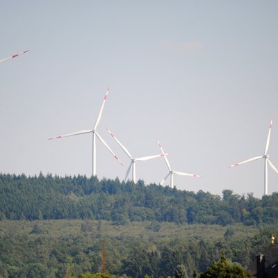 Auf dem Bild sind im Hintergrund mehrere Windräder zu sehen, welche sich vom Himmel abheben. Im Vordergrund sieht man einen Kirchturm.